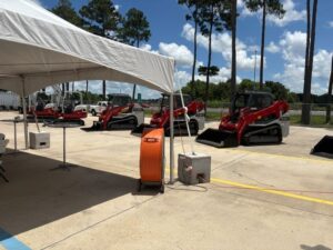 Outdoor equipment display under a white canopy: an orange extension cord reel and a concrete power box sit in the foreground while three red skid-steer loaders are lined up on a sunny concrete lot with trees in the background.