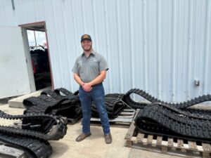 Man in work shirt and cap standing outdoors beside piles of rubber tank tracks on pallets outside a metal building.